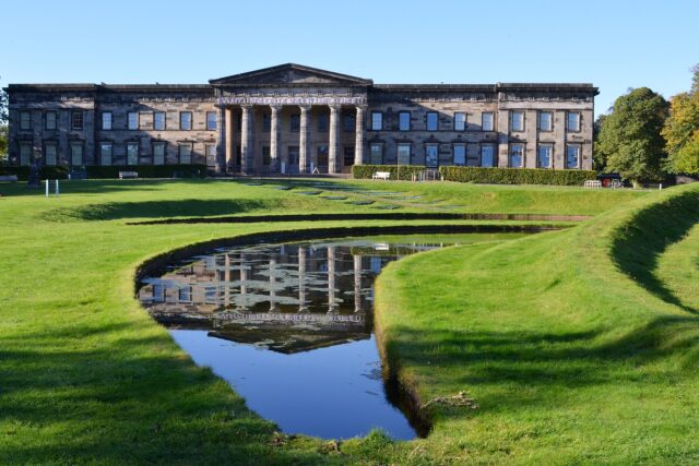 Modern One in Edinburgh overlooks the curved grassy banks and reflective pool of Landform under a clear blue sky.