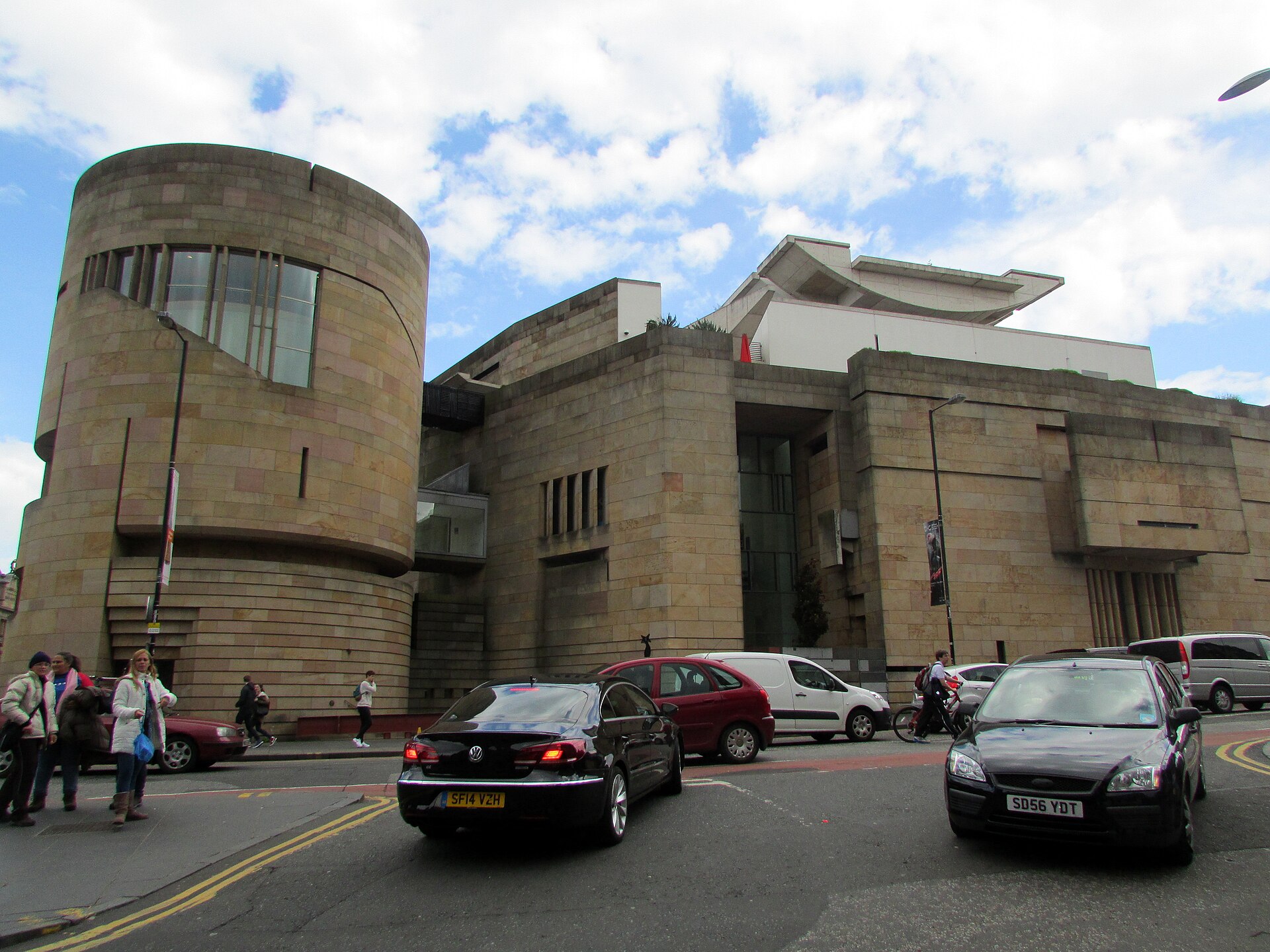 The National Museum of Scotland sandstone exterior and cylindrical tower overlook pedestrians and traffic on Chambers Street in Edinburgh.