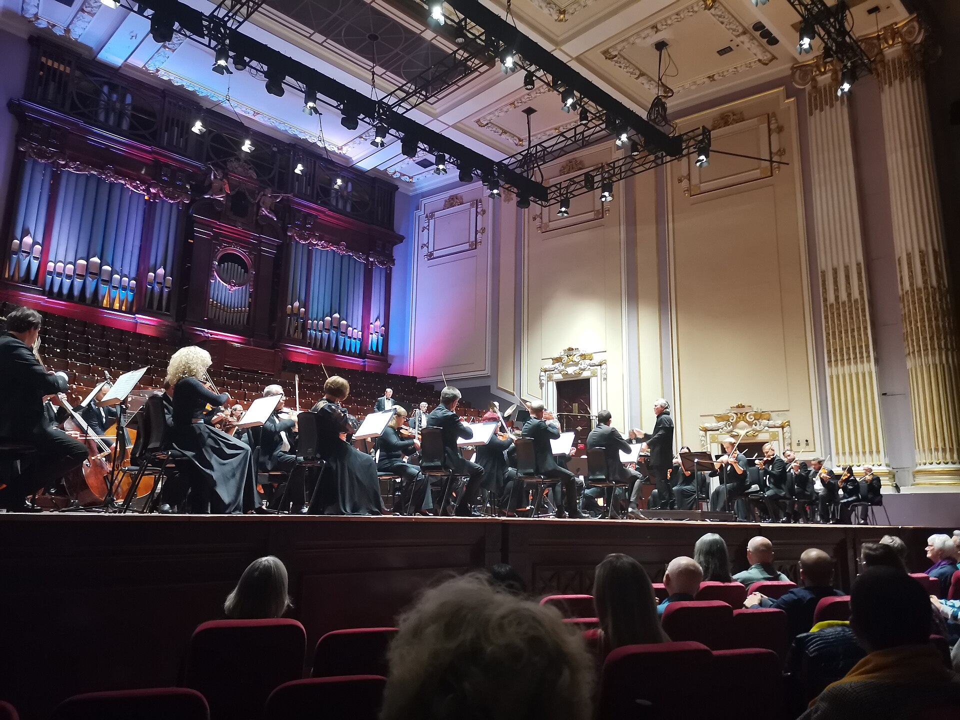An orchestra performs beneath the pipe organ at Edinburgh's Usher Hall, with the conductor and seated audience visible.