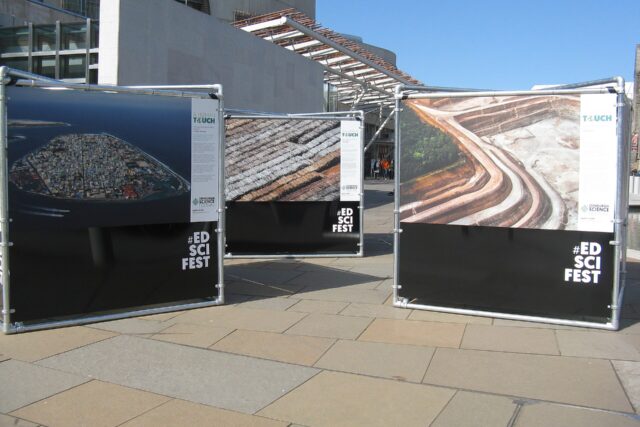 Outdoor Edinburgh Science Festival exhibition panels stand beside the Scottish Parliament beneath a clear blue sky.