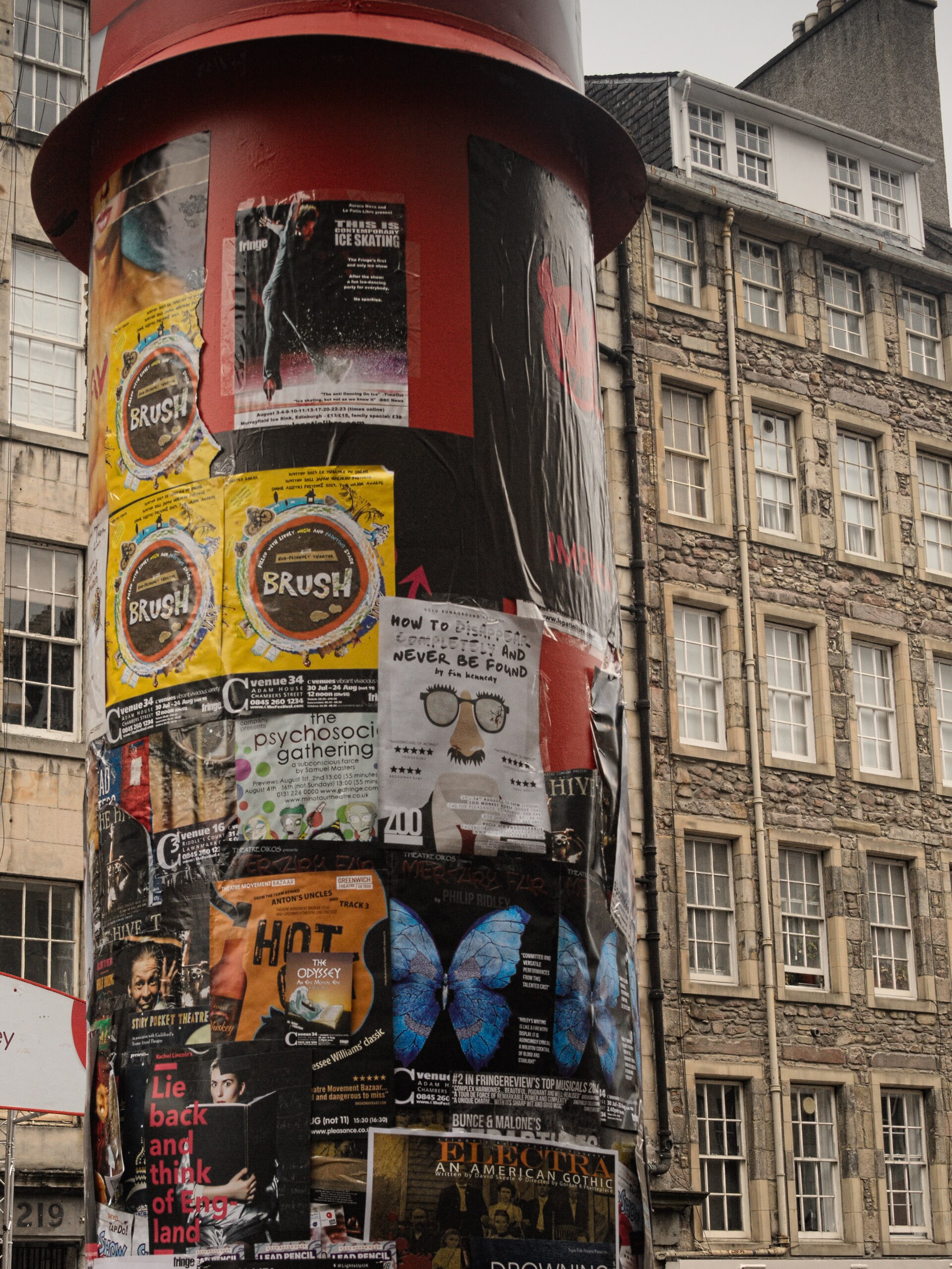 Overlapping Fringe show posters cover a red advertising column on Edinburgh's High Street, with tall stone buildings behind it.