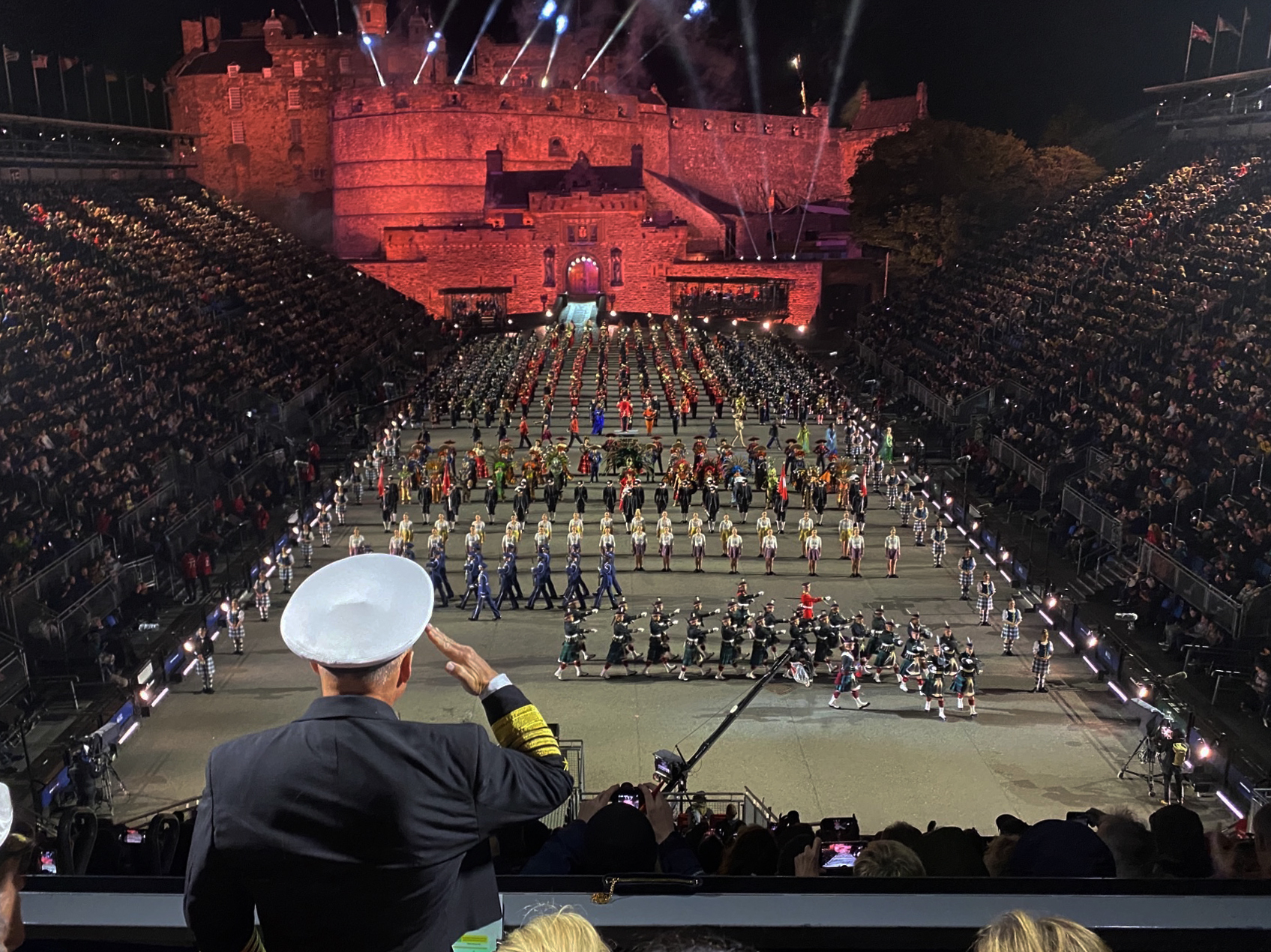 Packed grandstands flank Edinburgh Castle Esplanade during the Military Tattoo, with massed performers and a saluting officer in the foreground.