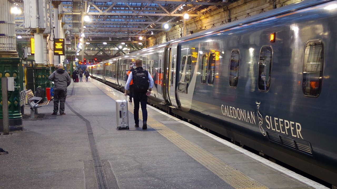 Passengers with luggage stand beside teal Caledonian Sleeper coaches beneath the roof at Edinburgh Waverley station.