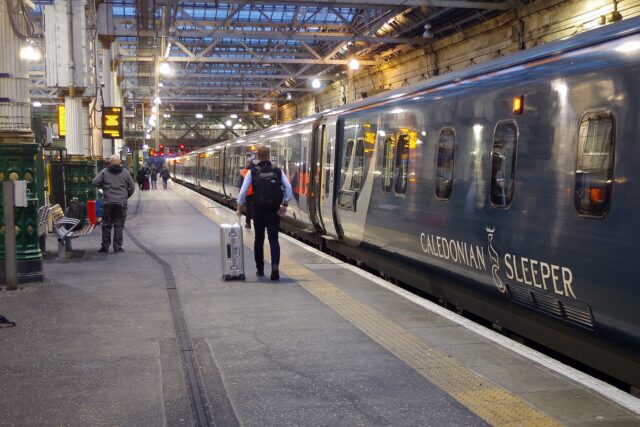 Passengers with luggage stand beside teal Caledonian Sleeper coaches beneath the roof at Edinburgh Waverley station.
