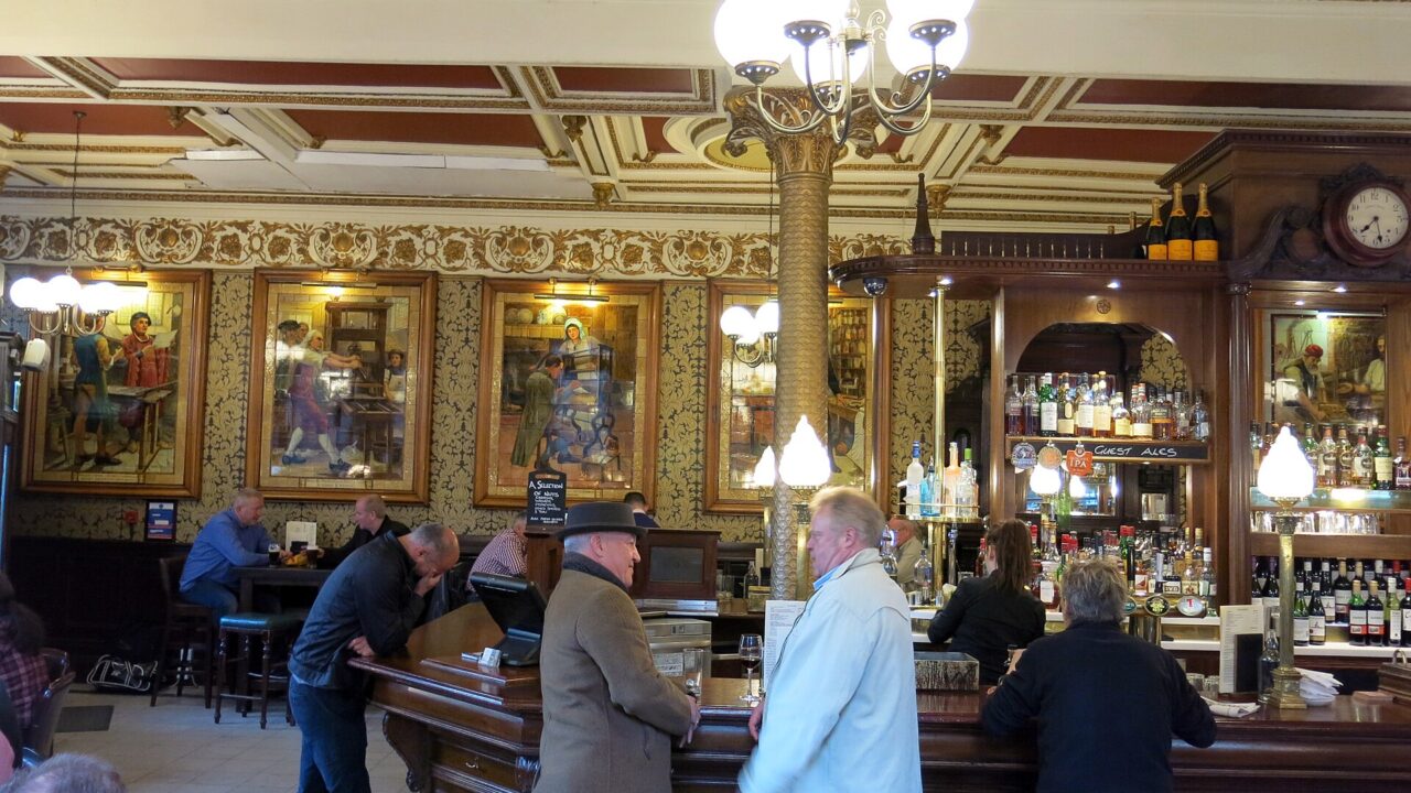Patrons gather around the curved wooden bar at Edinburgh's Café Royal beneath gilded ceiling mouldings and decorative tiled panels.