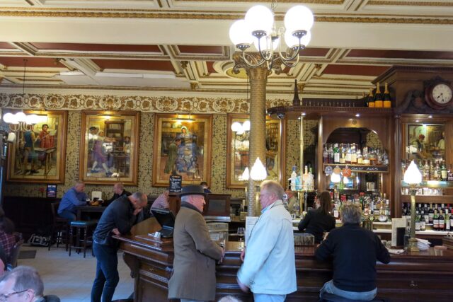Patrons gather around the curved wooden bar at Edinburgh's Café Royal beneath gilded ceiling mouldings and decorative tiled panels.