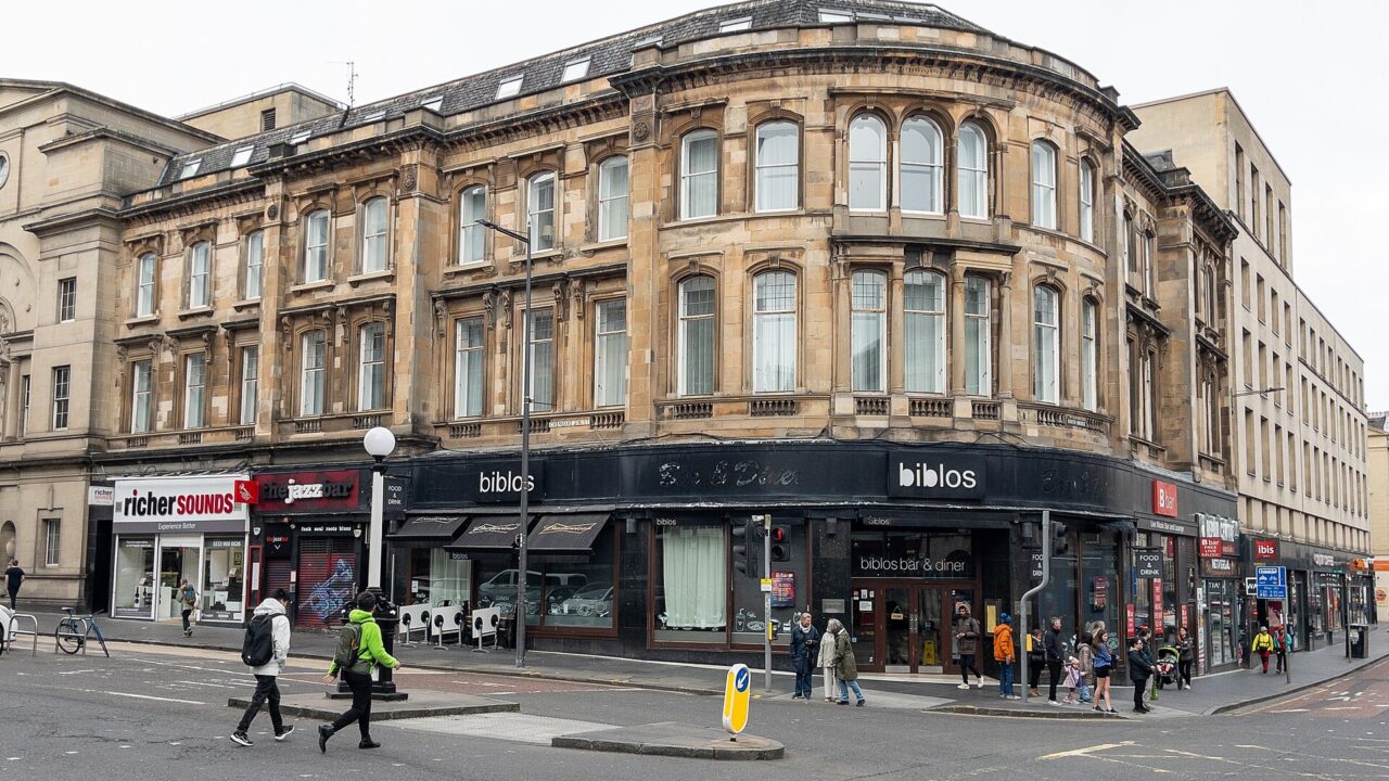 Pedestrians cross South Bridge in Edinburgh beside a curved stone building with shops and restaurants at street level.