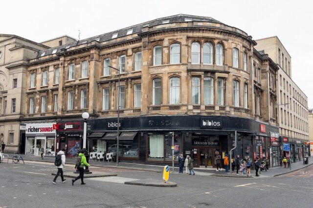 Pedestrians cross South Bridge in Edinburgh beside a curved stone building with shops and restaurants at street level.