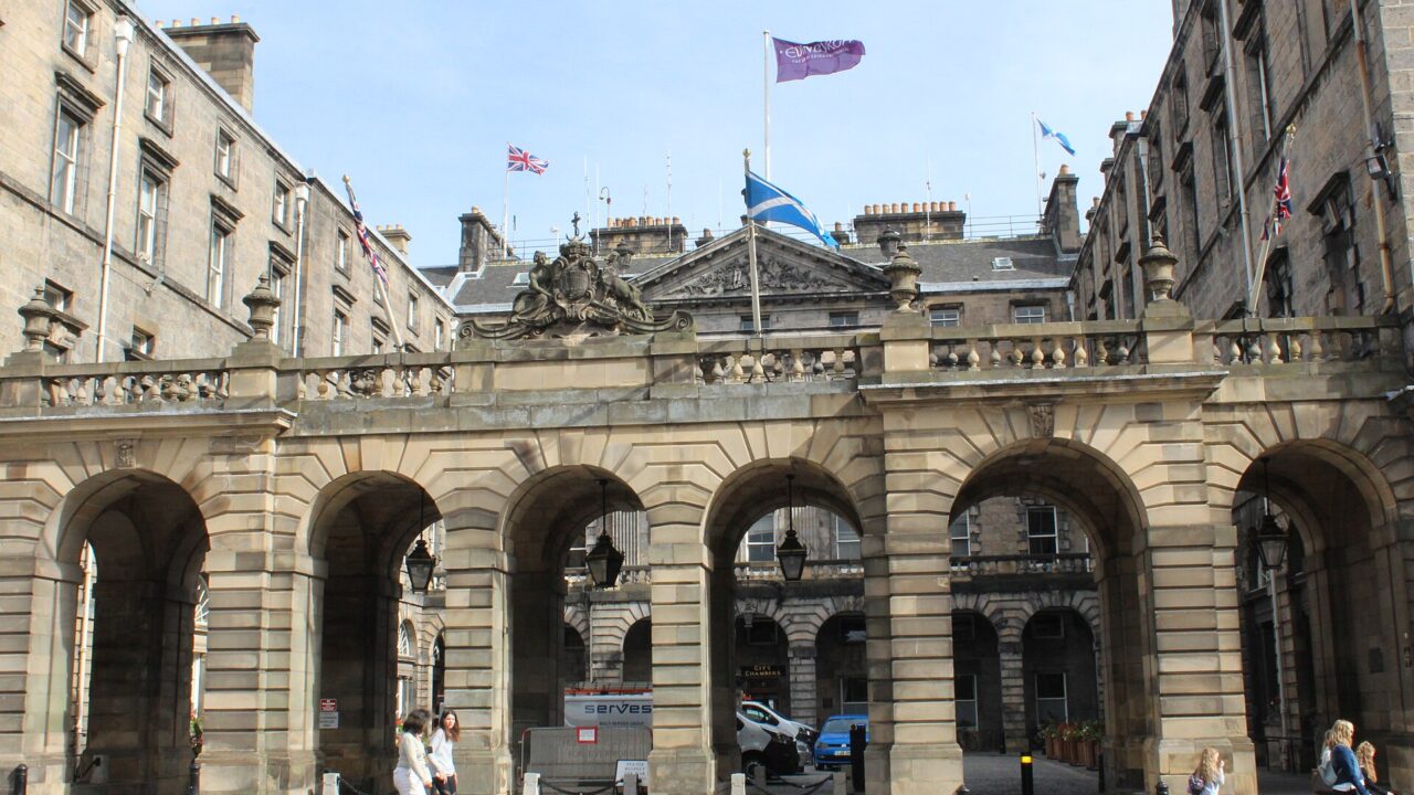 Pedestrians pass the stone arcade of Edinburgh City Chambers on the Royal Mile, with flags flying above the courtyard buildings.
