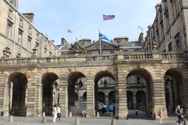 Pedestrians pass the stone arcade of Edinburgh City Chambers on the Royal Mile, with flags flying above the courtyard buildings.