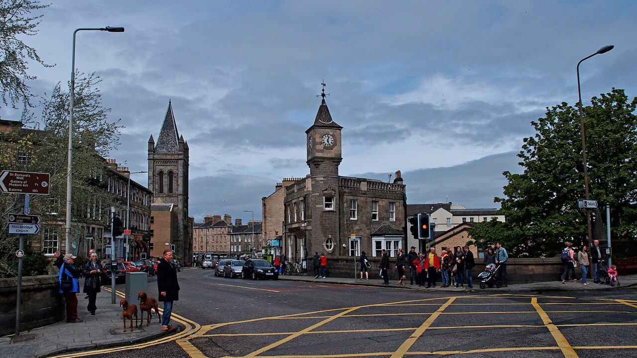 Pedestrians and traffic cross Stockbridge in Edinburgh beside a stone clock-towered building and a church spire.