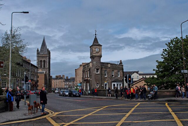 Pedestrians and traffic cross Stockbridge in Edinburgh beside a stone clock-towered building and a church spire.