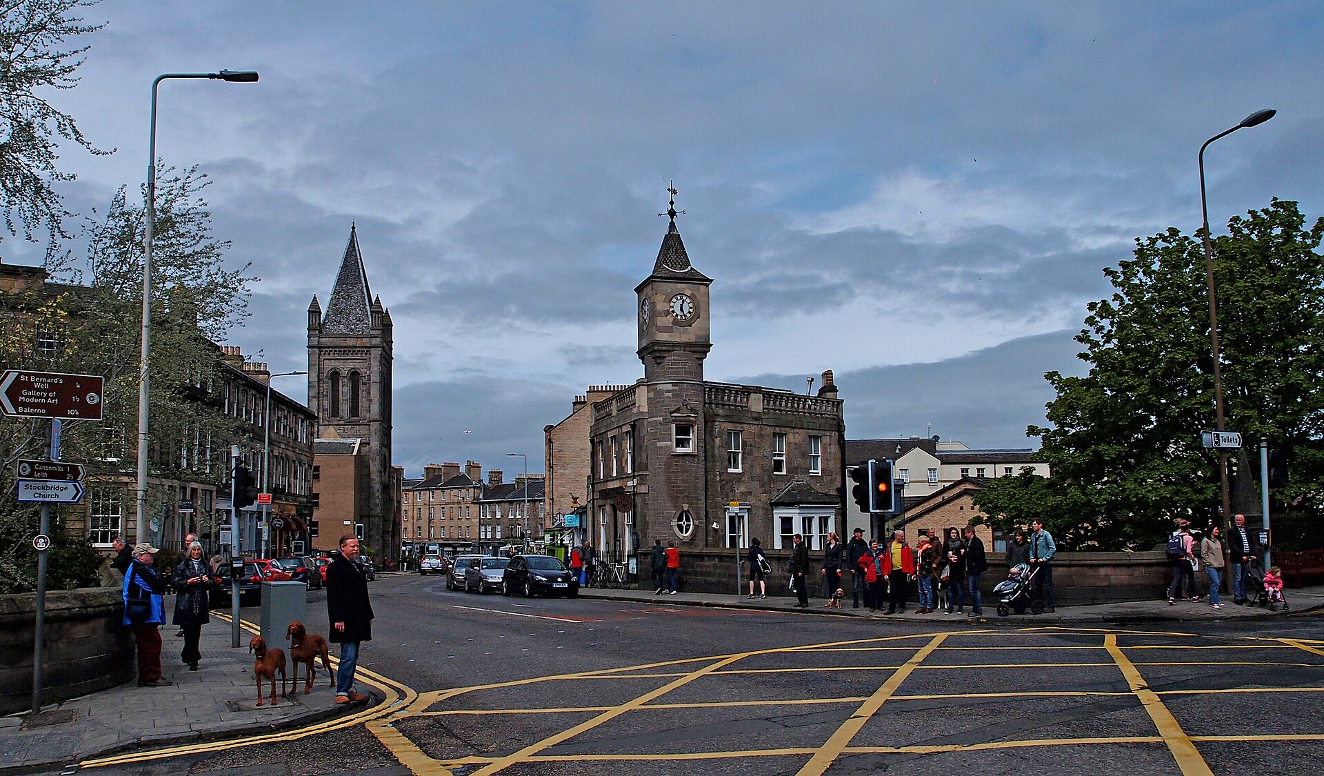 Pedestrians and traffic cross Stockbridge in Edinburgh beside a stone clock-towered building and a church spire.