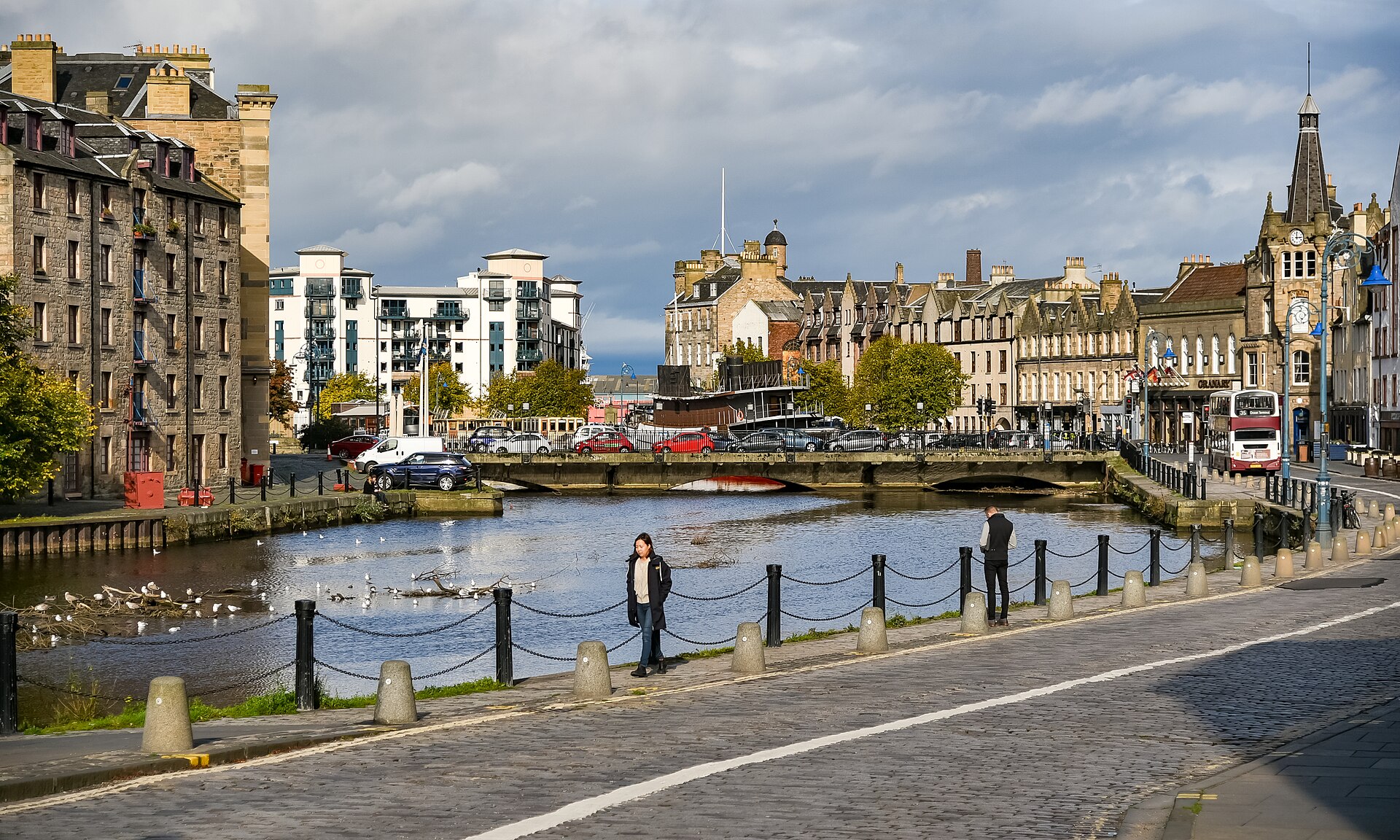 Pedestrians walk along the chain-lined Water of Leith quayside, with a low bridge and waterfront buildings ahead.