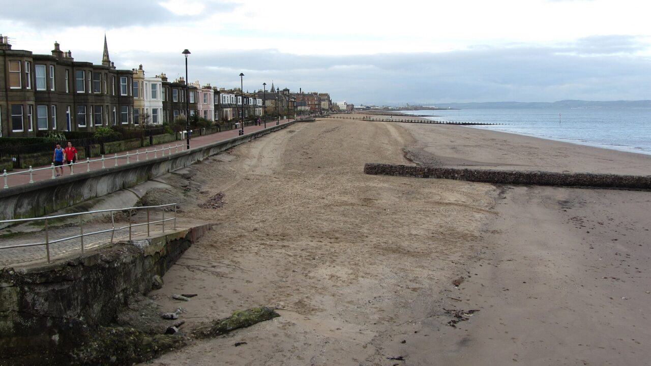 Portobello Beach stretches beside Edinburgh seafront houses and a promenade, with timber groynes extending toward the Firth of Forth.