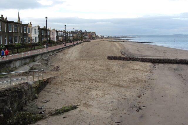 Portobello Beach stretches beside Edinburgh seafront houses and a promenade, with timber groynes extending toward the Firth of Forth.
