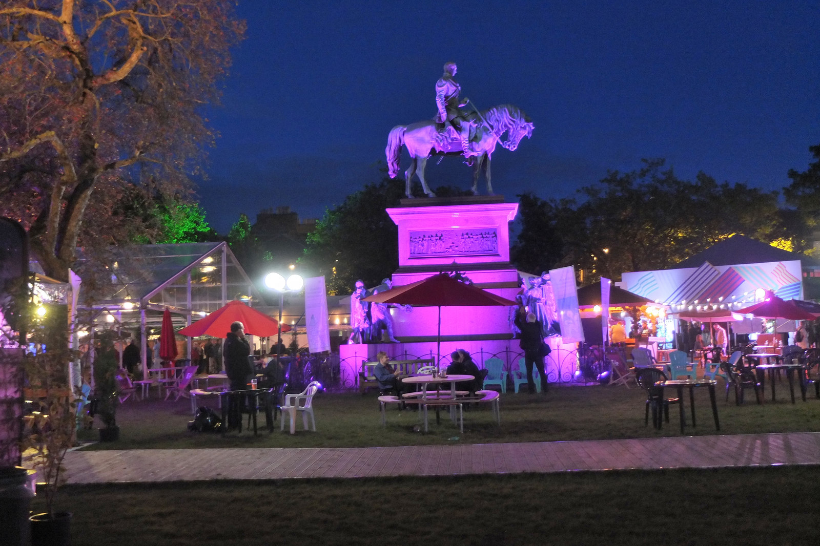 Purple lighting illuminates Charlotte Square's equestrian statue above tents and outdoor tables at the Edinburgh International Book Festival after dark.