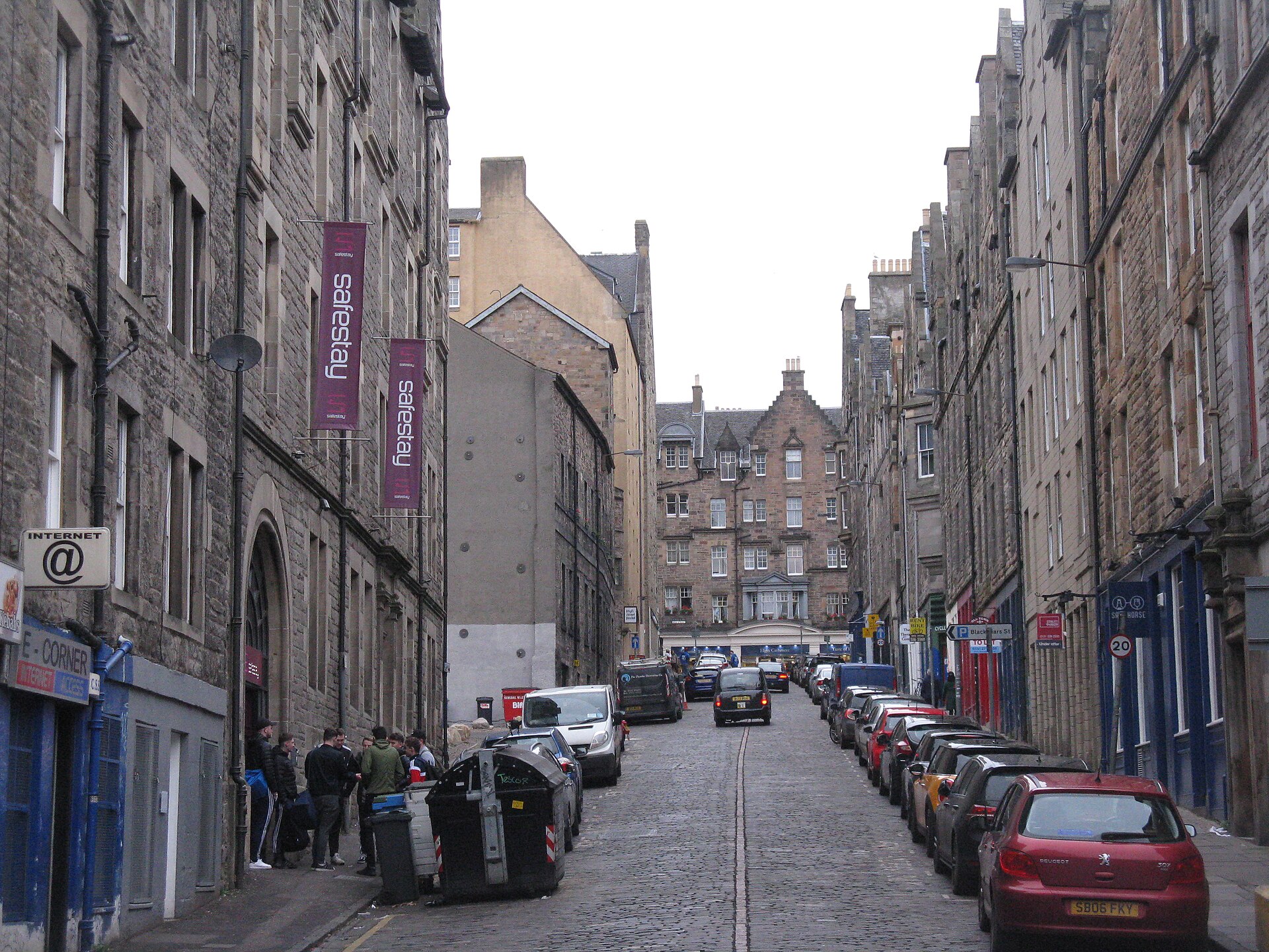 Purple Safestay signs hang above people outside the hostel on cobbled Blackfriars Street in Edinburgh, with parked cars lining the road.