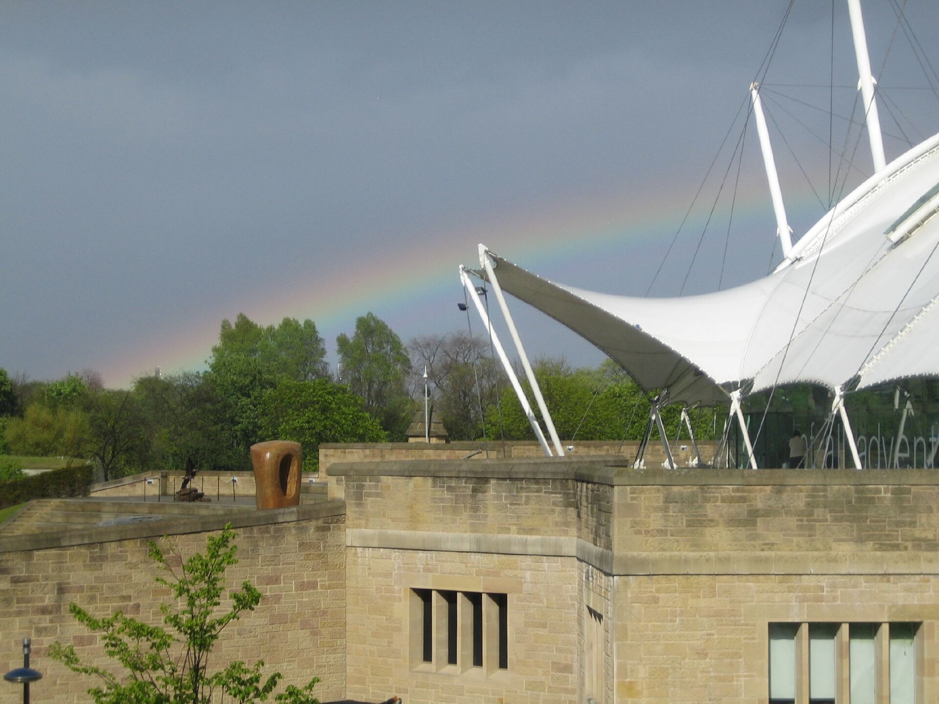 A rainbow stretches behind Dynamic Earth in Edinburgh, above green trees and the building's white roof and stone walls.