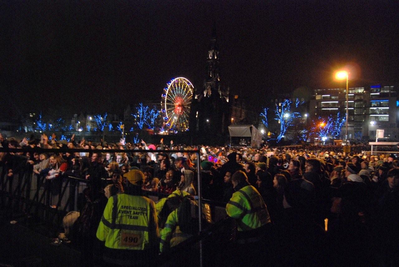 Revellers crowd Edinburgh's Hogmanay street party beside barriers, with the Scott Monument and a brightly lit Ferris wheel behind them.