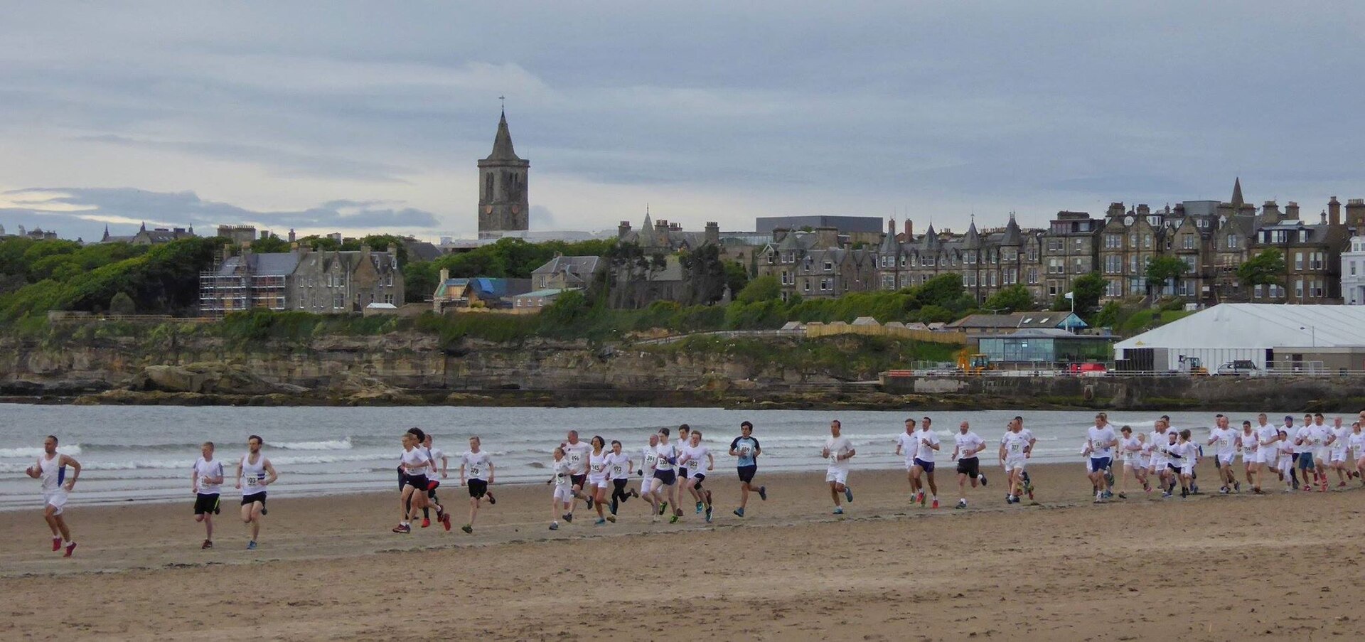 Runners cross West Sands at St Andrews, with breaking waves, stone buildings and a church tower behind them.