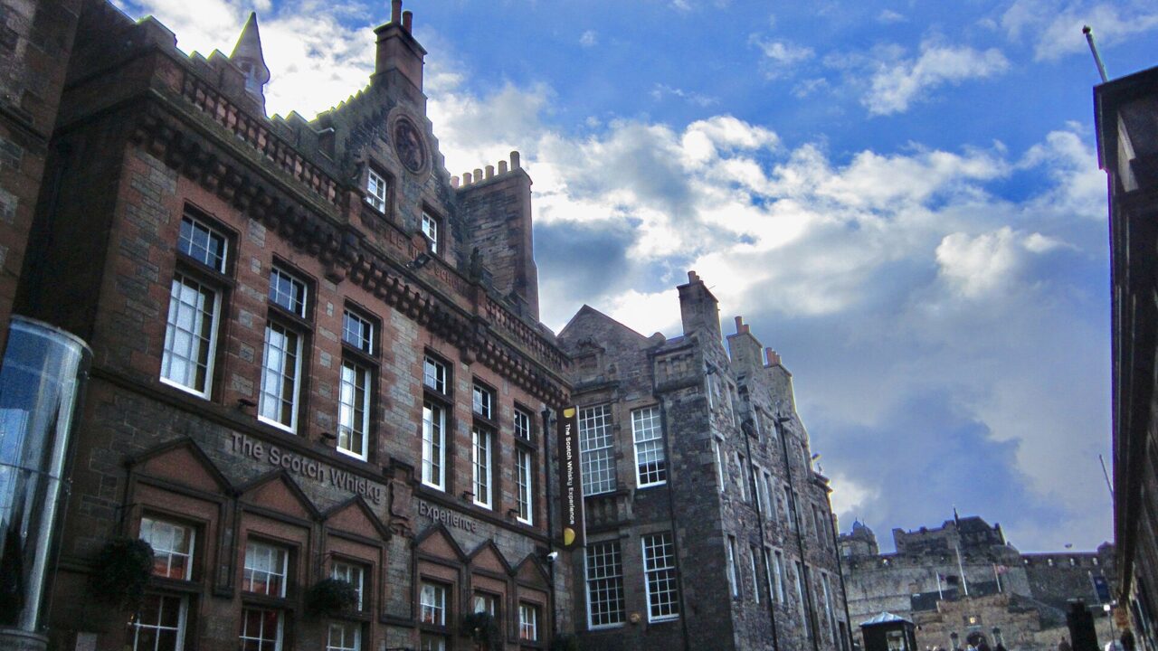 The Scotch Whisky Experience's red sandstone frontage lines Edinburgh's Castlehill, with pedestrians and Edinburgh Castle visible further uphill.