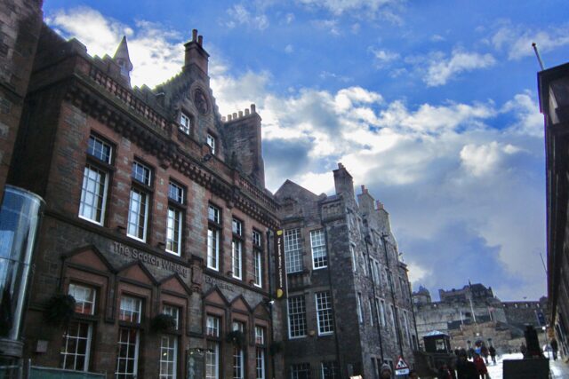 The Scotch Whisky Experience's red sandstone frontage lines Edinburgh's Castlehill, with pedestrians and Edinburgh Castle visible further uphill.