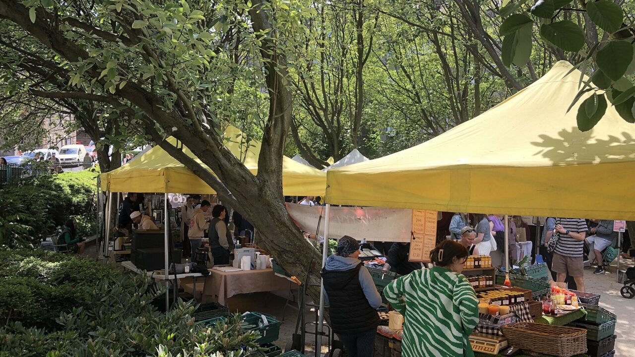 Shoppers browse jars and bottles beneath yellow canopies among the trees at Edinburgh's Stockbridge Market on a sunny day.