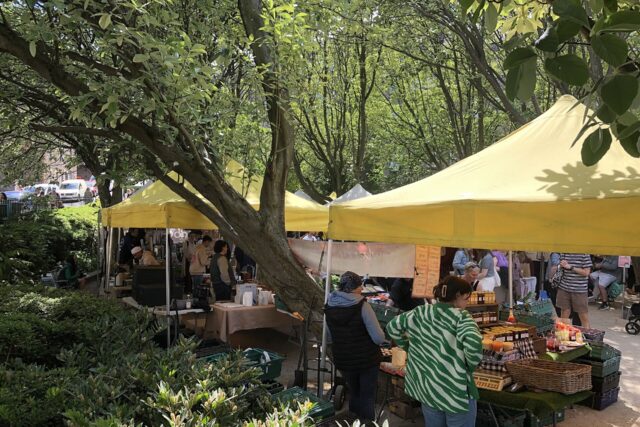Shoppers browse jars and bottles beneath yellow canopies among the trees at Edinburgh's Stockbridge Market on a sunny day.