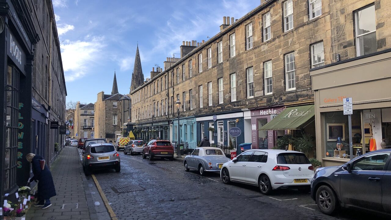 Shops line cobbled William Street in Edinburgh, with stone tenements above and church spires beyond the parked cars.