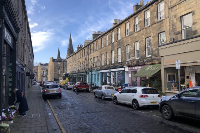 Shops line cobbled William Street in Edinburgh, with stone tenements above and church spires beyond the parked cars.