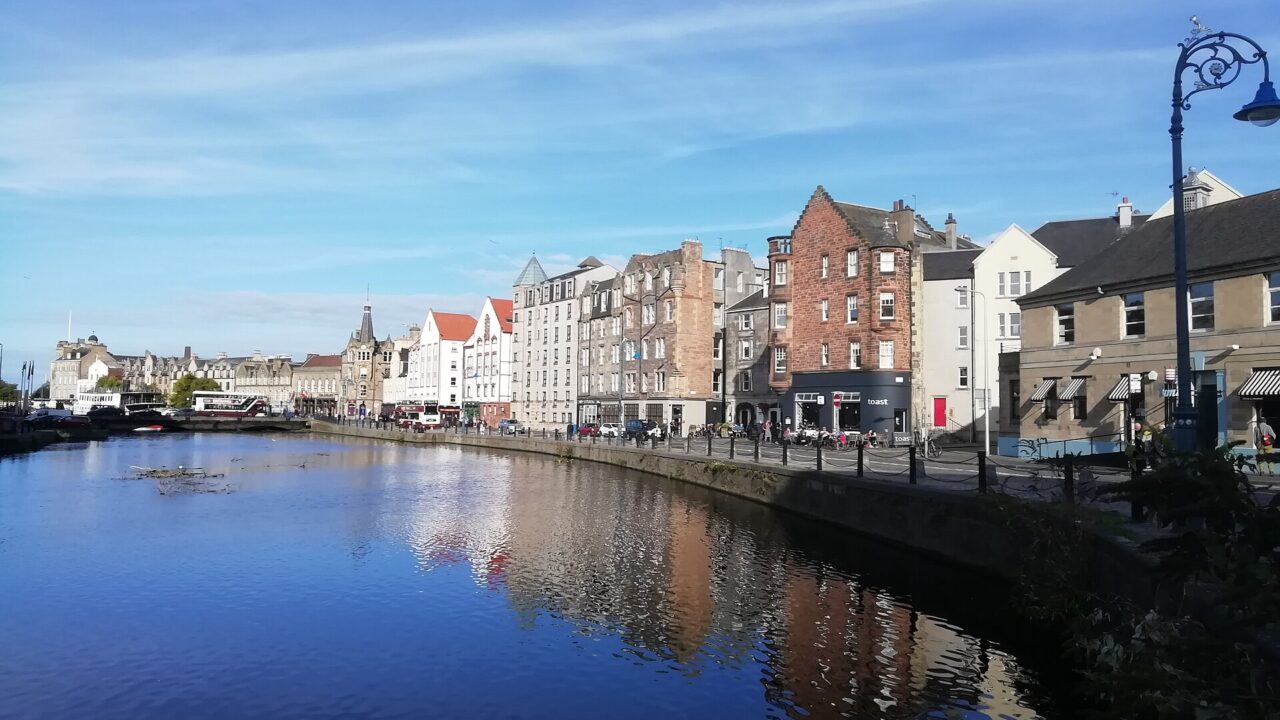 The Shore's stone and brick waterfront buildings reflect in the Water of Leith beneath a blue Edinburgh sky.