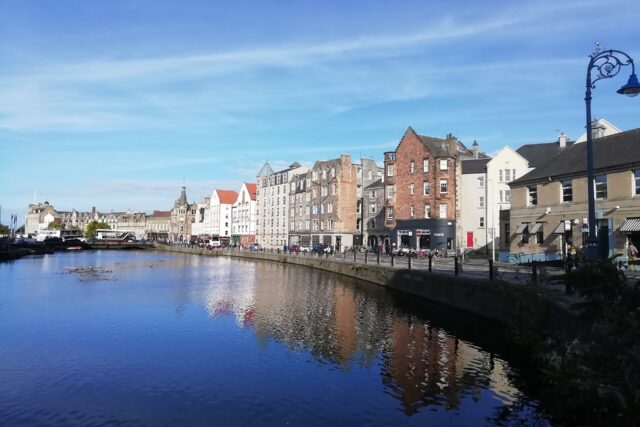 The Shore's stone and brick waterfront buildings reflect in the Water of Leith beneath a blue Edinburgh sky.