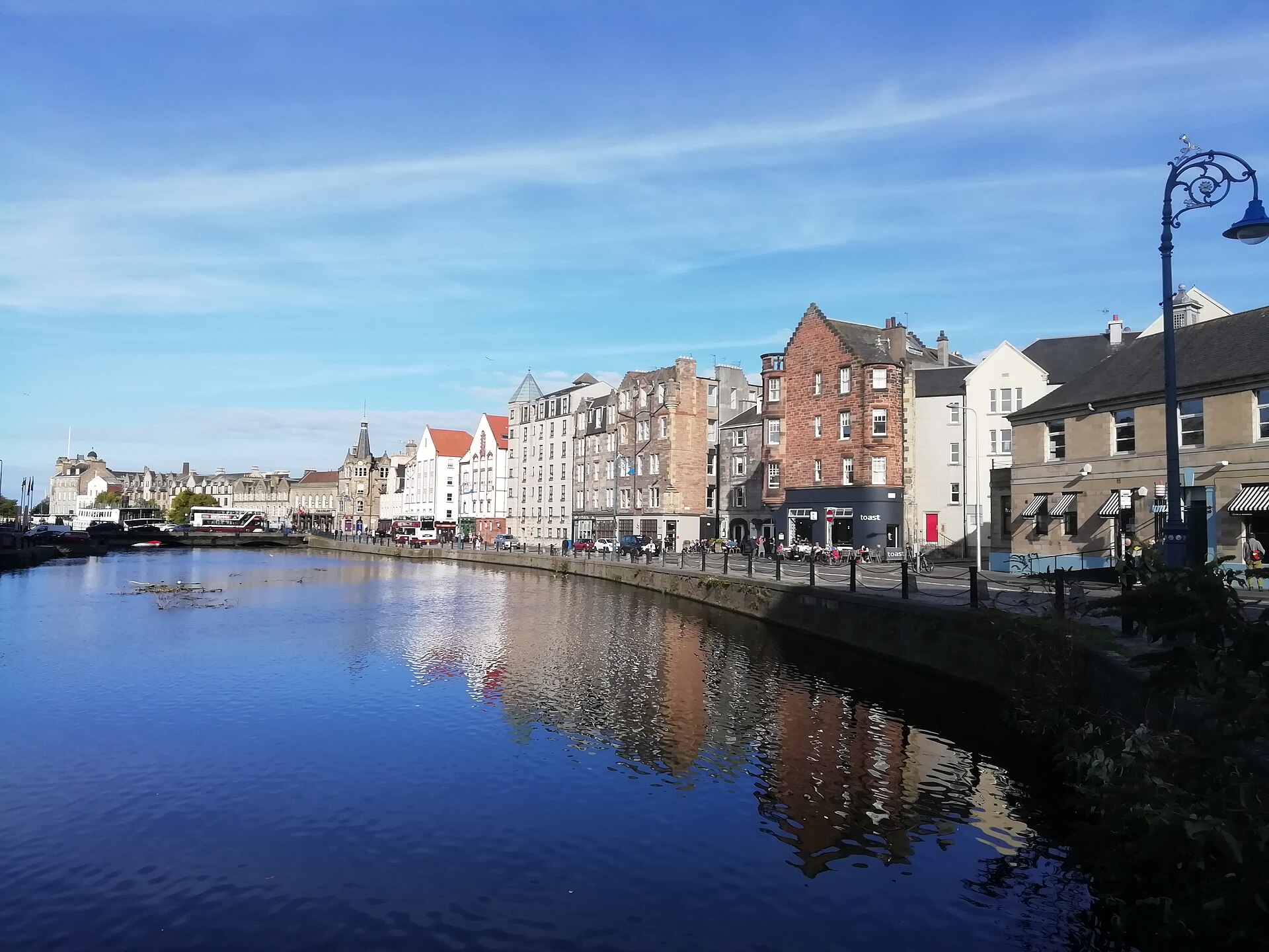 The Shore's stone and brick waterfront buildings reflect in the Water of Leith beneath a blue Edinburgh sky.