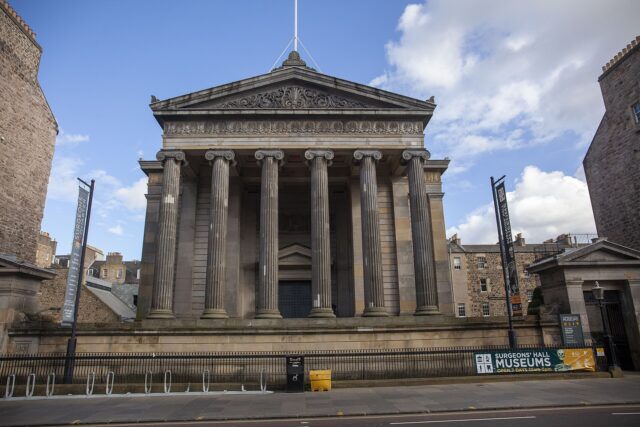 The six Ionic columns and carved pediment of Surgeons' Hall in Edinburgh rise behind railings and museum banners.