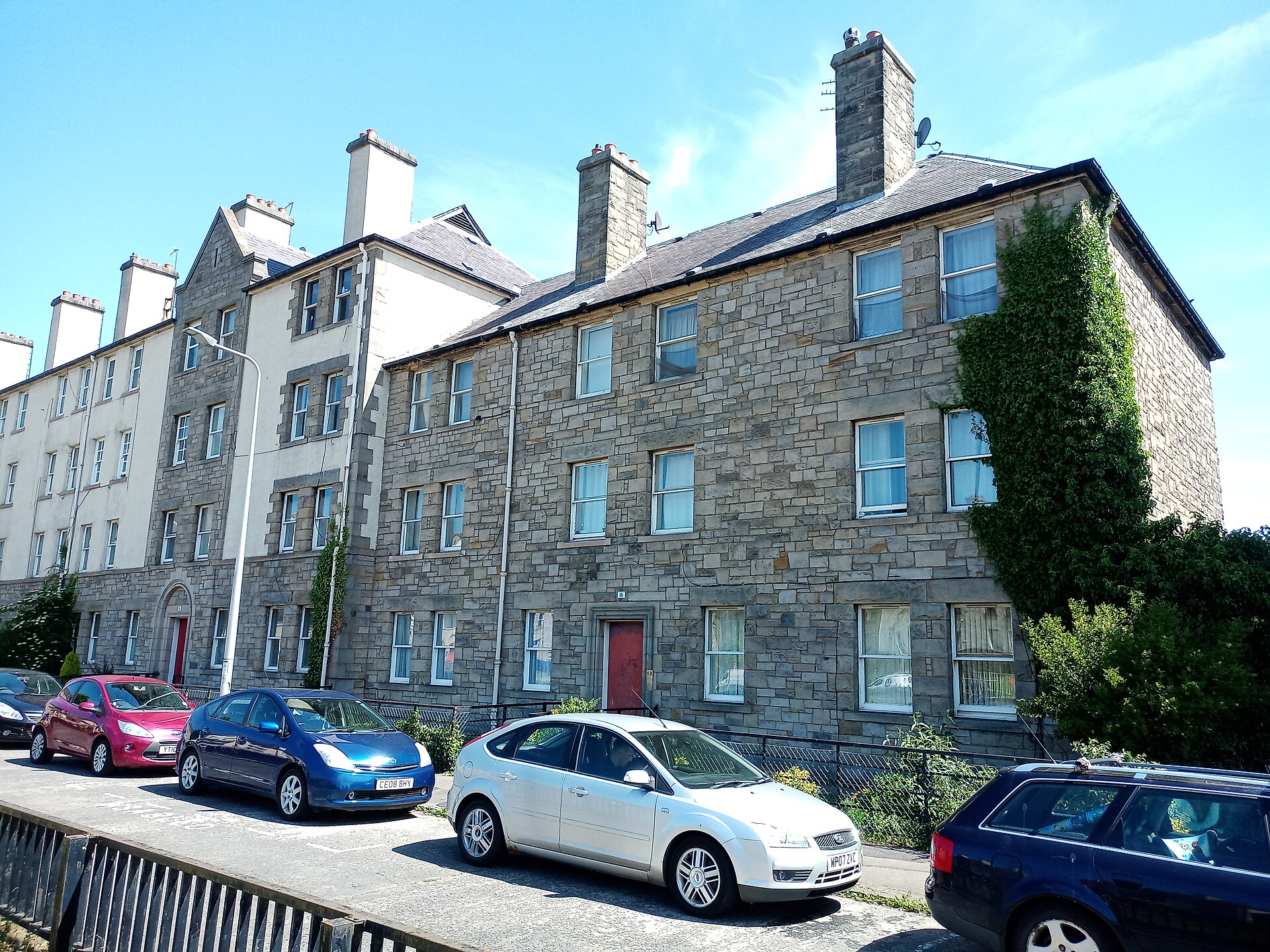 Stone tenements at Piershill Square West in Edinburgh have red entrance doors, tall chimneys and ivy beside parked cars.
