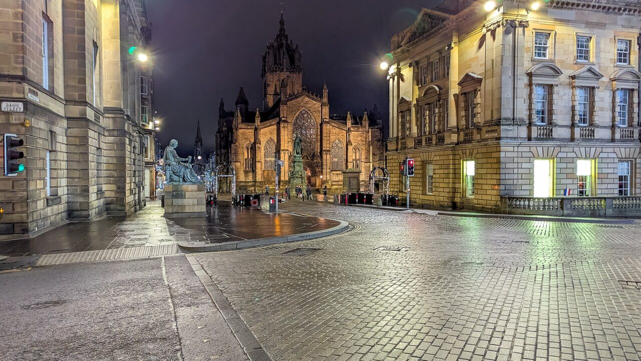 Streetlights illuminate wet cobbles in Edinburgh Old Town at night, with St Giles Cathedral framed by stone buildings.