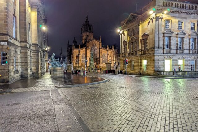 Streetlights illuminate wet cobbles in Edinburgh Old Town at night, with St Giles Cathedral framed by stone buildings.