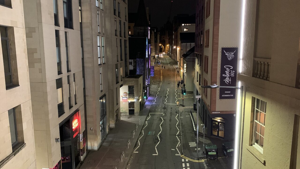 Streetlights and illuminated bar frontages line Edinburgh's Cowgate at night, with pedestrians walking between tall stone buildings.