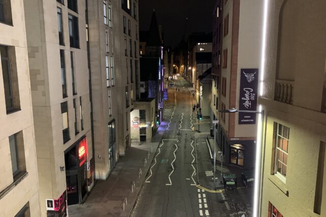 Streetlights and illuminated bar frontages line Edinburgh's Cowgate at night, with pedestrians walking between tall stone buildings.