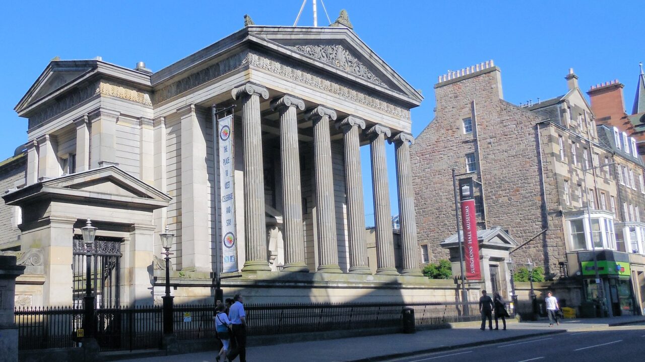 Surgeons’ Hall in Edinburgh presents its Ionic portico to Nicolson Street, with pedestrians passing the railings in sunshine.