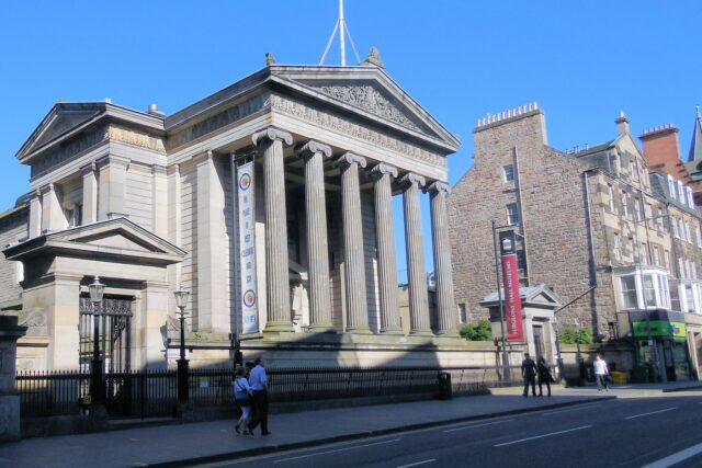 Surgeons’ Hall in Edinburgh presents its Ionic portico to Nicolson Street, with pedestrians passing the railings in sunshine.