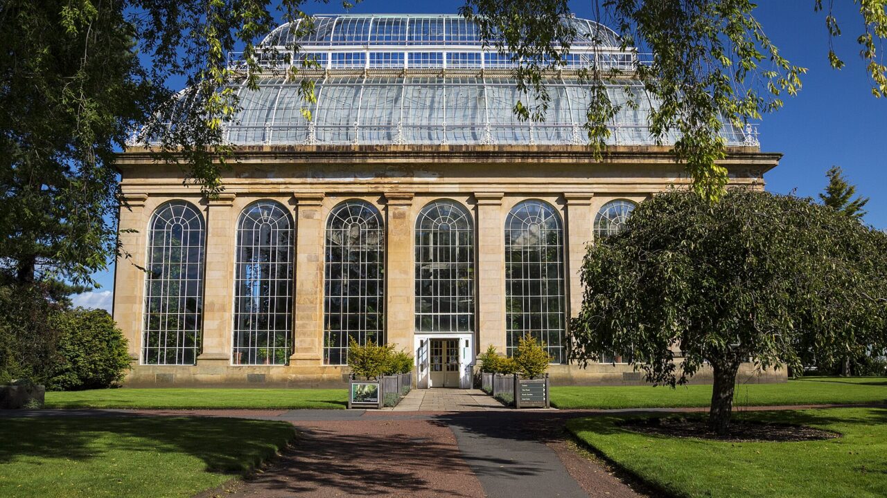 Tall arched windows and a glass roof crown the Palm House beside lawns at the Royal Botanic Garden Edinburgh.