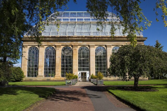 Tall arched windows and a glass roof crown the Palm House beside lawns at the Royal Botanic Garden Edinburgh.