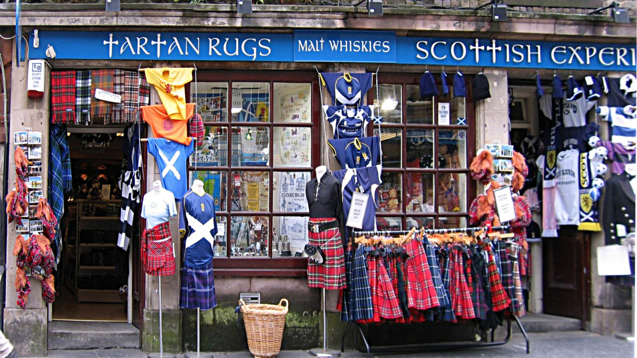 Tartan kilts hang outside a Royal Mile shop in Edinburgh, alongside flags, shirts and souvenirs beneath a blue fascia.