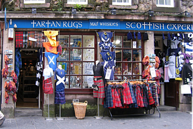 Tartan kilts hang outside a Royal Mile shop in Edinburgh, alongside flags, shirts and souvenirs beneath a blue fascia.