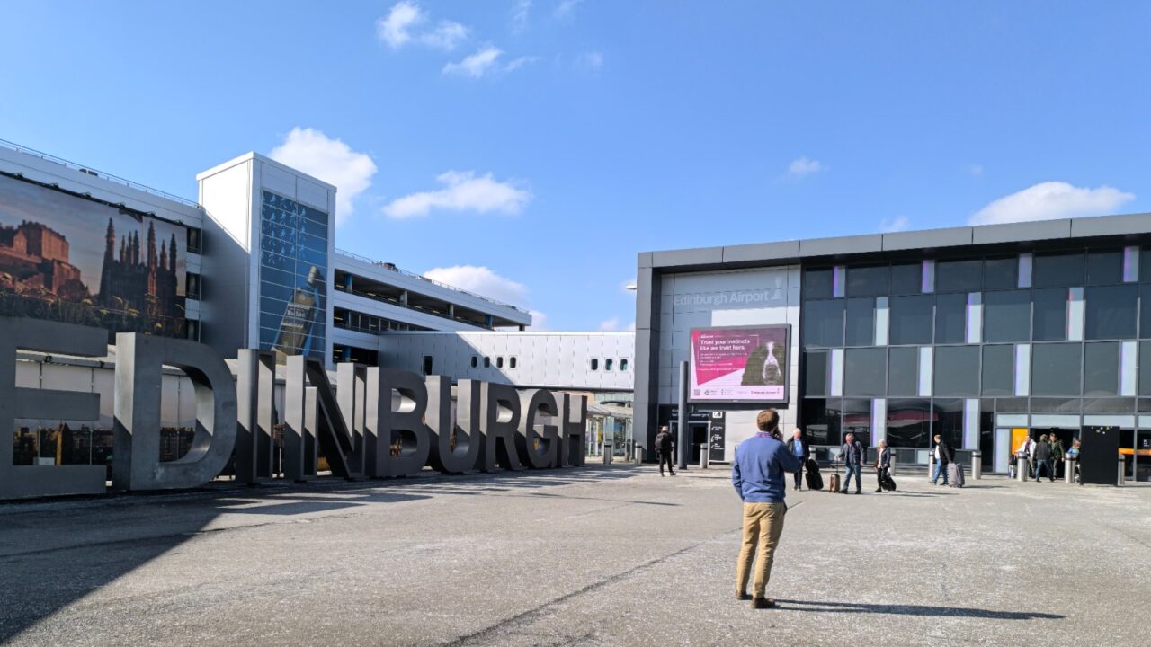 Travellers cross the forecourt outside Edinburgh Airport beside giant Edinburgh letters and the glass-fronted terminal entrance.
