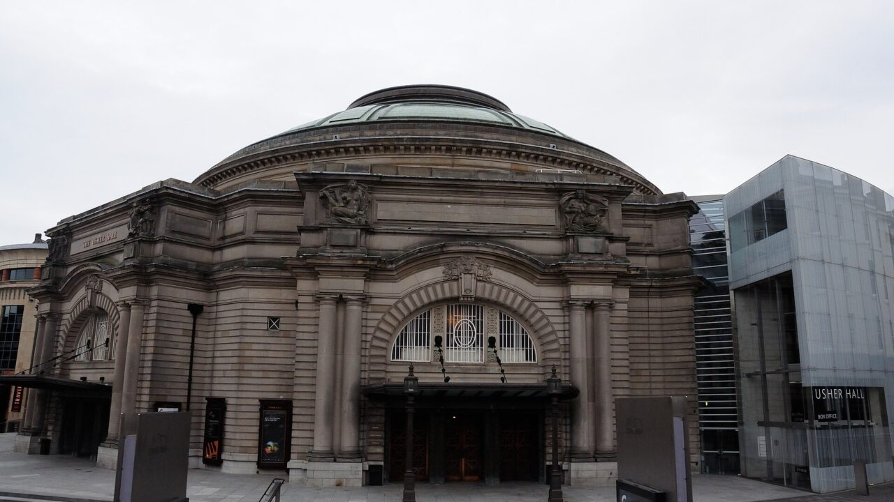 Usher Hall in Edinburgh has an arched stone entrance beneath a broad dome, beside a modern glass extension.