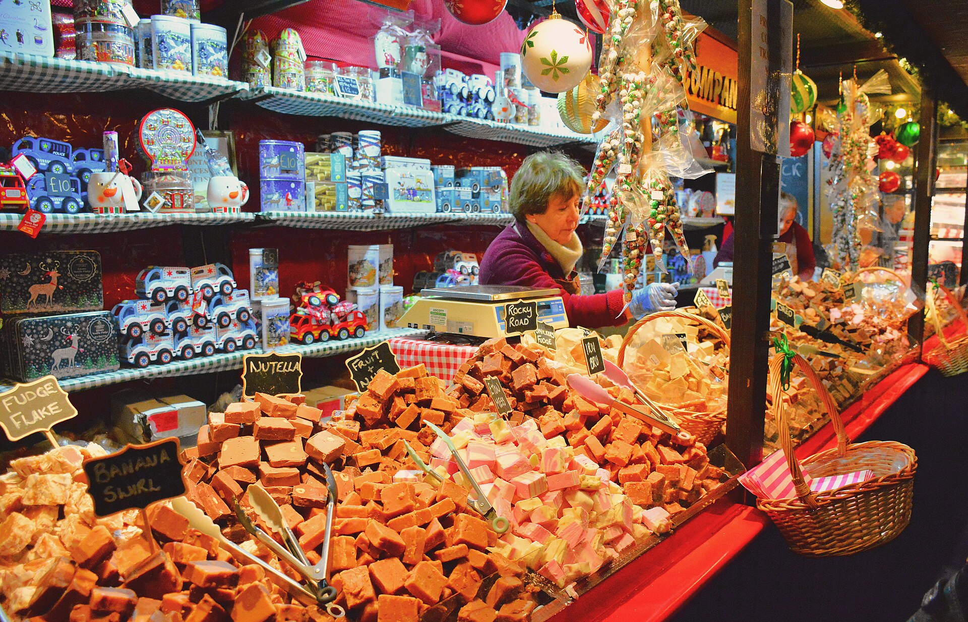 A vendor serves behind trays of fudge and other sweets at Edinburgh Christmas Market, surrounded by gift tins and decorations.