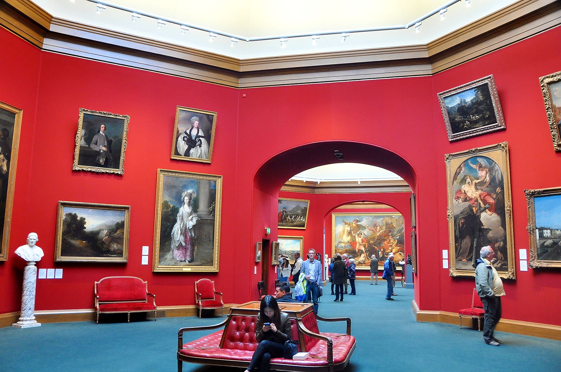 Visitors browse paintings or rest on red upholstered seats beneath broad arches in Edinburgh's Scottish National Gallery.